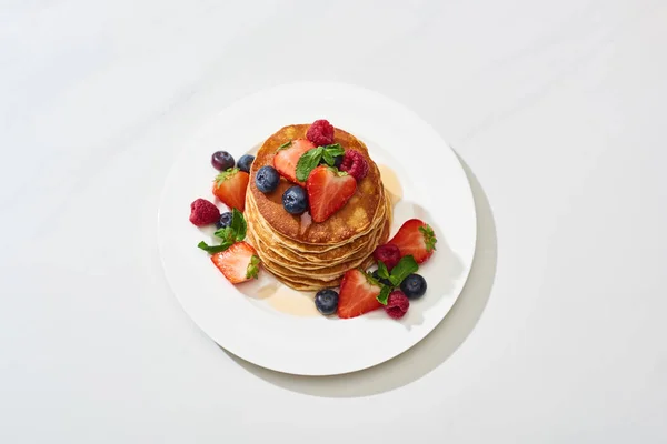 Blick von oben auf leckere Pfannkuchen mit Honig, Blaubeeren und Erdbeeren auf Teller auf weißem Marmor — Stockfoto