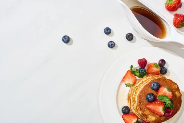 Blick von oben auf leckere Pfannkuchen mit Ahornsirup, Blaubeeren und Erdbeeren auf Teller auf weißem Marmor — Stockfoto
