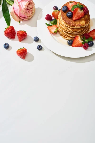 Vista superior de deliciosas panquecas com mirtilos e morangos na placa perto de flor de rosa na superfície branca de mármore — Fotografia de Stock