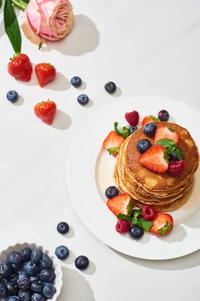 Blick von oben auf leckere Pfannkuchen mit Blaubeeren und Erdbeeren auf Teller in der Nähe von Rosenblüte auf weißem Marmor — Stockfoto