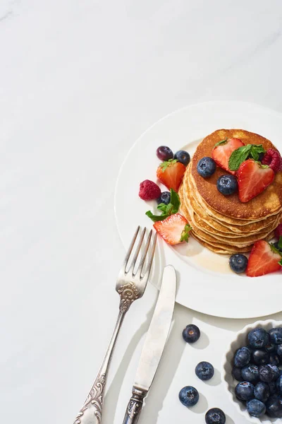 Top view of delicious pancakes with maple syrup, blueberries and strawberries on plate near fork and knife on marble white surface — Stock Photo