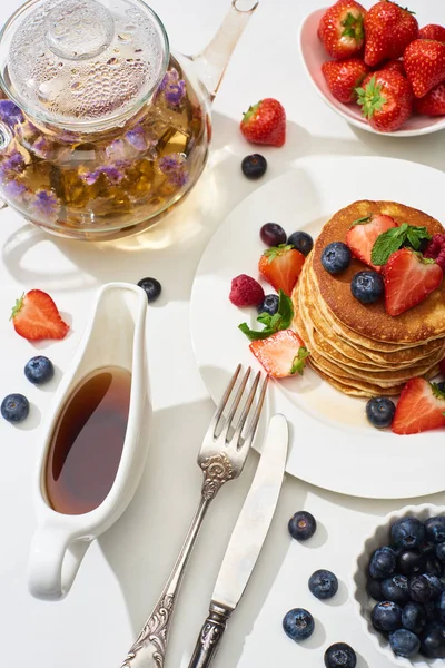 Top view of delicious pancakes with maple syrup, blueberries and strawberries on plate near herbal tea in teapot, fork and knife on marble white surface — Stock Photo