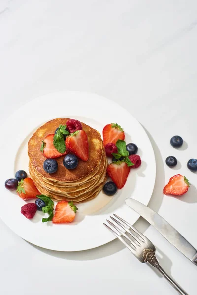 Top view of delicious pancakes with maple syrup, blueberries and strawberries on plate near fork and knife on marble white surface — Stock Photo