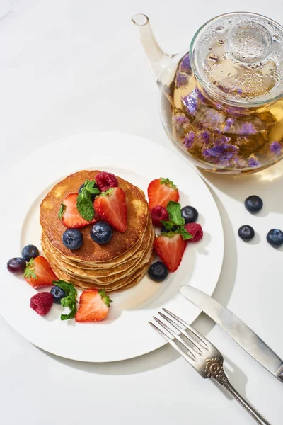 Top view of delicious pancakes with maple syrup, blueberries and strawberries on plate near herbal tea in teapot, fork and knife on marble white surface — Stock Photo