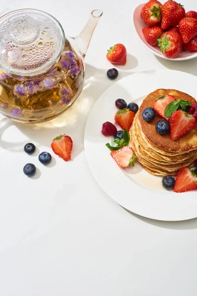 Top view of delicious pancakes with maple syrup, blueberries and strawberries on plate near herbal tea in teapot on marble white surface — Stock Photo
