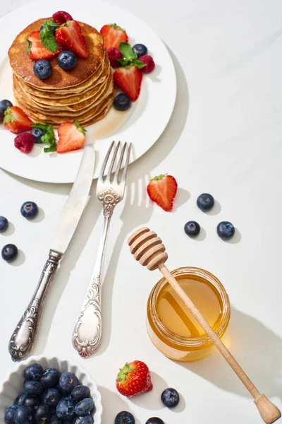 Vue de dessus des crêpes délicieuses avec du miel, des bleuets et des fraises sur la plaque près de la fourchette et couteau sur la surface de marbre blanc — Photo de stock