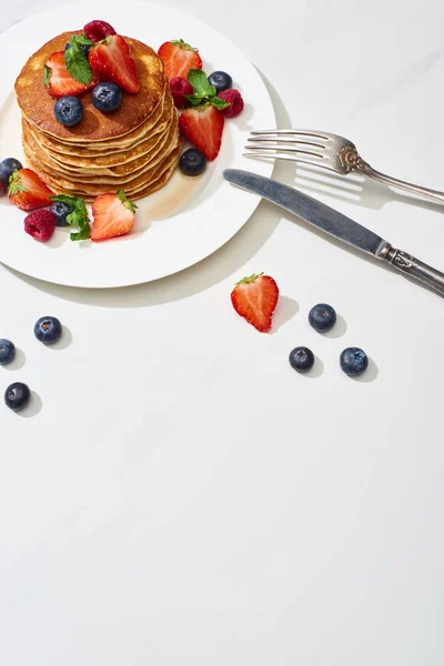 Top view of delicious pancakes with maple syrup, blueberries and strawberries on plate near fork and knife on marble white surface — Stock Photo