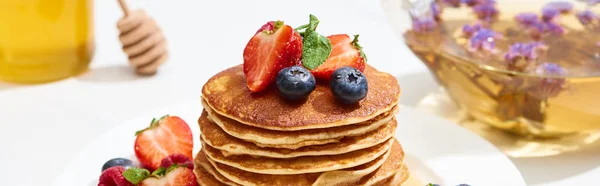 Mise au point sélective de délicieuses crêpes au miel, aux bleuets et aux fraises sur assiette à la surface blanche, prise de vue panoramique — Photo de stock