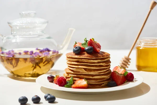 Selective focus of delicious pancakes with honey, blueberries and strawberries on plate near herbal tea on white surface isolated on grey — Stock Photo