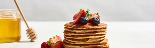 Selektiver Fokus von leckeren Pfannkuchen mit Blaubeeren und Erdbeeren in der Nähe von Honig auf weißer Oberfläche isoliert auf grauer, panoramischer Aufnahme — Stockfoto