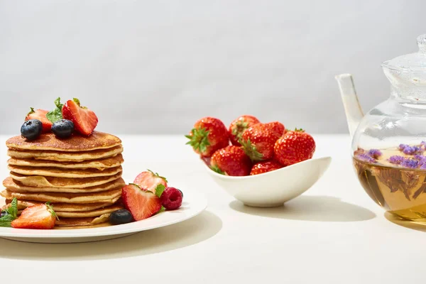 Leckere Pfannkuchen mit Blaubeeren und Erdbeeren auf Teller in der Nähe von Kräutertee in Teekanne auf weißer Oberfläche isoliert auf grau — Stockfoto