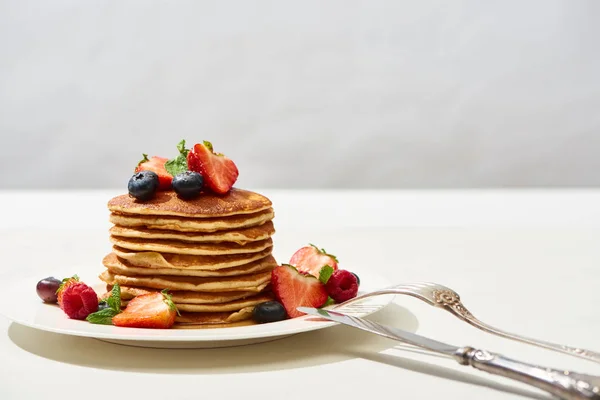 Deliziosi pancake con mirtilli e fragole su piatto con posate su superficie bianca isolata su grigio — Foto stock