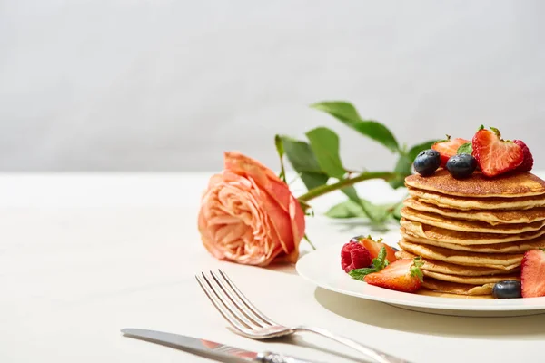 Selective focus of delicious pancakes with blueberries and strawberries on plate near rose flower and cutlery on white surface isolated on grey — Stock Photo