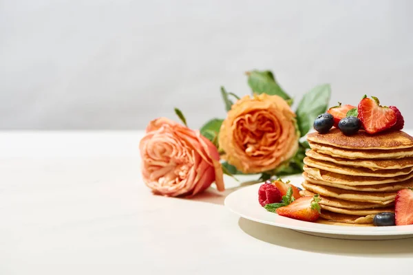 Foco seletivo de deliciosas panquecas com mirtilos e morangos na placa perto de flores de rosas na superfície branca isolada em cinza — Fotografia de Stock