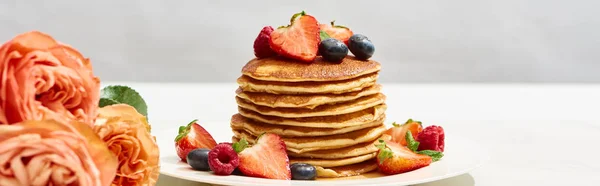 Selective focus of delicious pancakes with blueberries and strawberries on plate near rose flowers on white surface isolated on grey, panoramic shot — Stock Photo