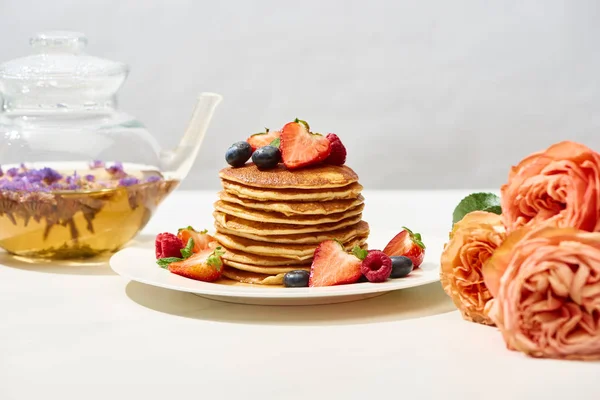 Selective focus of delicious pancakes with blueberries and strawberries on plate near rose flowers and herbal tea on white surface isolated on grey — Stock Photo