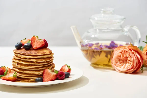 Foco seletivo de deliciosas panquecas com mirtilos e morangos no prato perto de flores de rosas e chá de ervas na superfície branca isolada em cinza — Fotografia de Stock