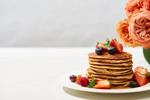 Deliciosos panqueques con arándanos y fresas en el plato cerca de flores de rosas en la superficie blanca aislada en gris - foto de stock