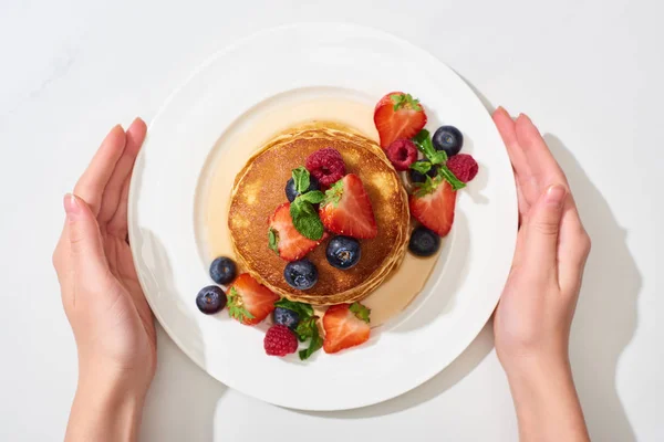 Partial view of woman holding plate with delicious pancakes with honey, blueberries and strawberries on marble white surface — Stock Photo