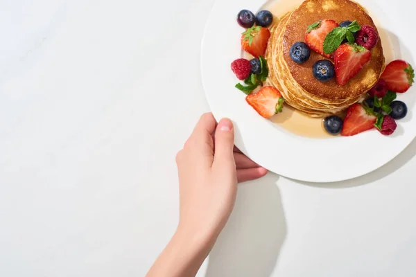 Частичный вид женщины, держащей тарелку с вкусными блинами с медом, черникой и клубникой на мраморной белой поверхности — стоковое фото