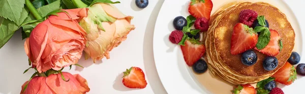 Top view of delicious pancakes with honey, blueberries and strawberries on plate near roses on marble white surface, panoramic shot — Stock Photo