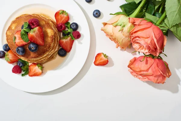 Top view of delicious pancakes with honey, blueberries and strawberries on plate near roses on marble white surface — Stock Photo