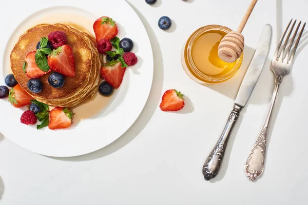 Top view of delicious pancakes with honey, blueberries and strawberries on plate near cutlery on marble white surface — Stock Photo