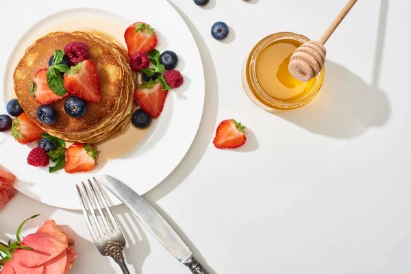 Top view of delicious pancakes with honey, blueberries and strawberries on plate near cutlery and rose on marble white surface — Stock Photo