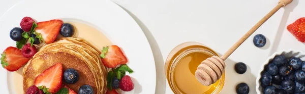 Vue de dessus des crêpes délicieuses avec du miel, des bleuets et des fraises sur plaque sur la surface de marbre blanc, vue panoramique — Photo de stock
