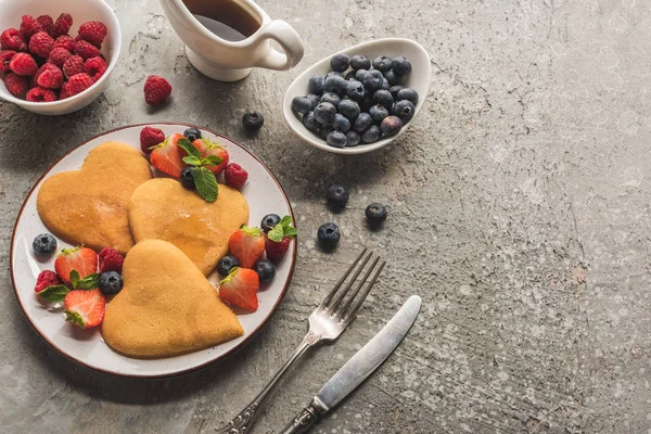 Herzförmige Pfannkuchen mit Beeren auf grauer Betonoberfläche mit Besteck und Ahornsirup — Stockfoto