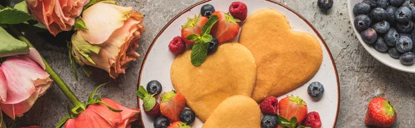 Draufsicht auf herzförmige Pfannkuchen mit Beeren auf grauer Betonoberfläche in der Nähe von Rosen, Panoramaaufnahme — Stockfoto