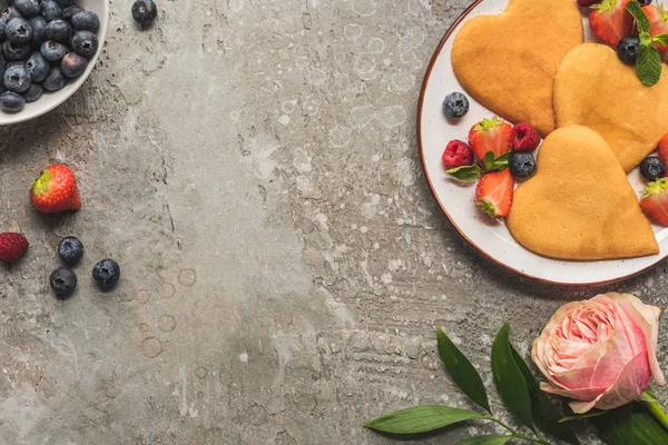 Top view of heart shaped pancakes with berries on grey concrete surface near rose — Stock Photo