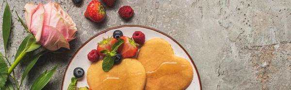 Top view of heart shaped pancakes with berries on grey concrete surface near rose, panoramic shot — Stock Photo