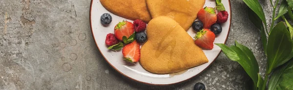 Top view of heart shaped pancakes with berries on grey concrete surface near plant, panoramic shot — Stock Photo