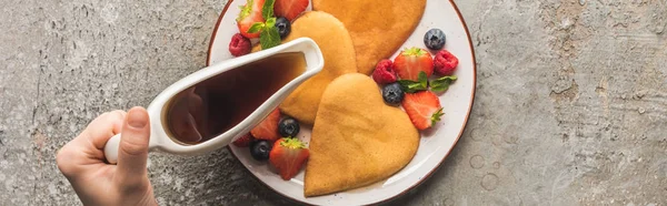 Vista ritagliata della donna che aggiunge sciroppo d'acero a frittelle a forma di cuore con bacche su una superficie di cemento grigio, colpo panoramico — Foto stock