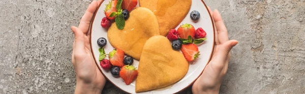 Cropped view of woman holding plate with heart shaped pancakes and berries on grey concrete surface, panoramic shot — Stock Photo