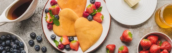 Top view of heart shaped pancakes with berries on grey concrete surface with butter, honey and maple syrup, panoramic shot — Stock Photo