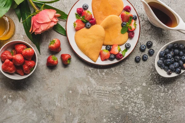 Vista dall'alto di frittelle a forma di cuore con bacche su una superficie di cemento grigio con rosa in fiore, miele e sciroppo d'acero — Foto stock