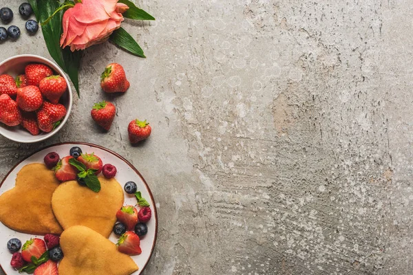 Vue de dessus des crêpes en forme de coeur avec des baies sur la surface en béton gris avec rose fleurie — Photo de stock