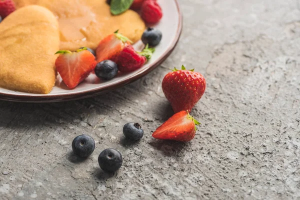 Vista de cerca de deliciosos panqueques en forma de corazón en el plato cerca de bayas en la superficie gris - foto de stock