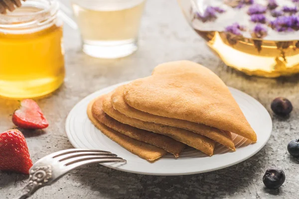 Foco seletivo de deliciosas panquecas em forma de coração com bagas e garfo perto de mel e chá de ervas na superfície de concreto cinza — Fotografia de Stock