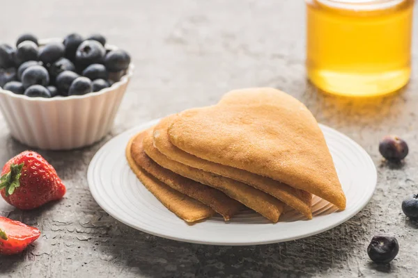 Délicieuses crêpes en forme de coeur avec des baies sur assiette près du miel sur surface en béton gris — Photo de stock