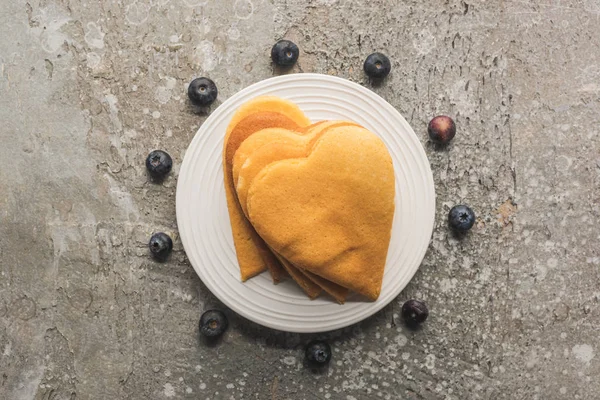 Top view of delicious heart shaped pancakes on plate near blueberries on grey concrete surface — Stock Photo