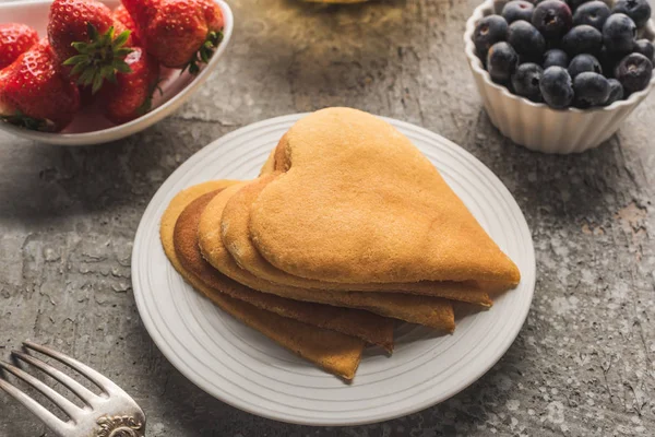 Deliciosas panquecas em forma de coração na placa com garfo perto de bagas na superfície de concreto cinza — Fotografia de Stock