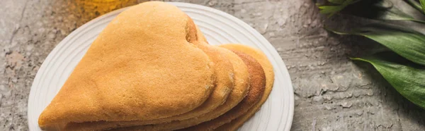 Delicious heart shaped pancakes on plate near plant on grey concrete surface, panoramic shot — Stock Photo