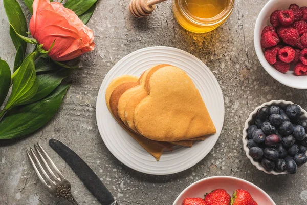 Vista dall'alto di frittelle a forma di cuore con bacche, miele, posate e rosa su una superficie di cemento grigio — Foto stock