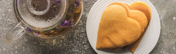 Top view of heart shaped pancakes near herbal tea in teapot on grey concrete surface, panoramic shot — Stock Photo