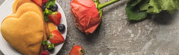 Вид зверху на млинці у формі серця зі свіжими ягодами на сірій бетонній поверхні з трояндою, панорамний знімок — стокове фото