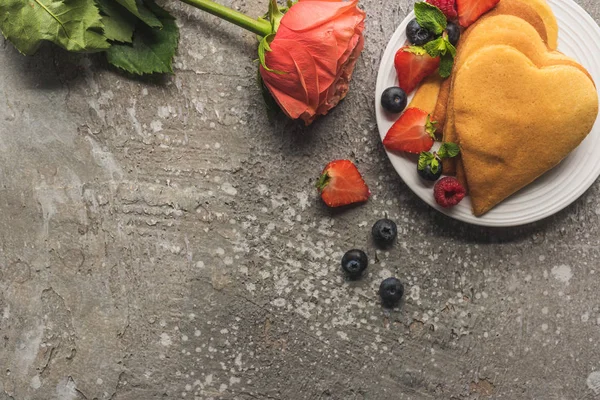 Vista dall'alto di frittelle a forma di cuore con bacche fresche su una superficie di cemento grigio con rosa — Foto stock