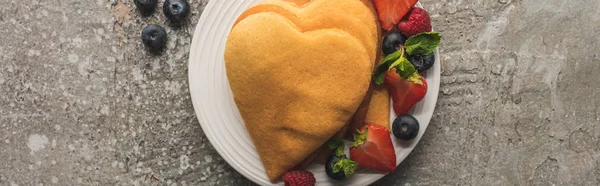 Top view of heart shaped pancakes with tasty berries on plate on grey concrete surface, panoramic shot — Stock Photo